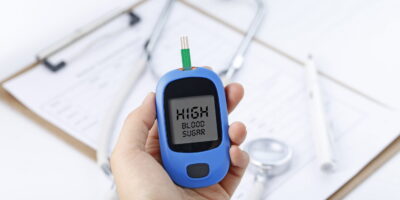 Hand holding a blood glucose meter measuring blood sugar, the background is a stethoscope and chart file